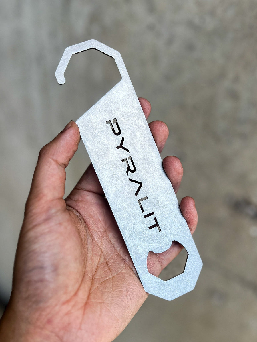 Metal bottle opener with 'PYRALIT' branding held in a hand against a neutral background